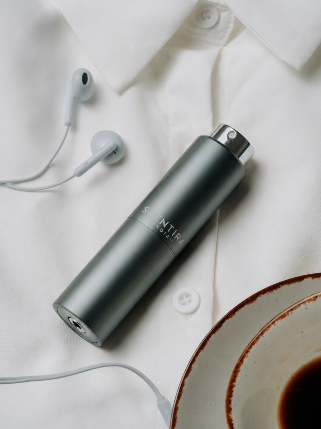 Scentira atomiser resting on a white shirt beside earphones and coffee — everyday premium carry